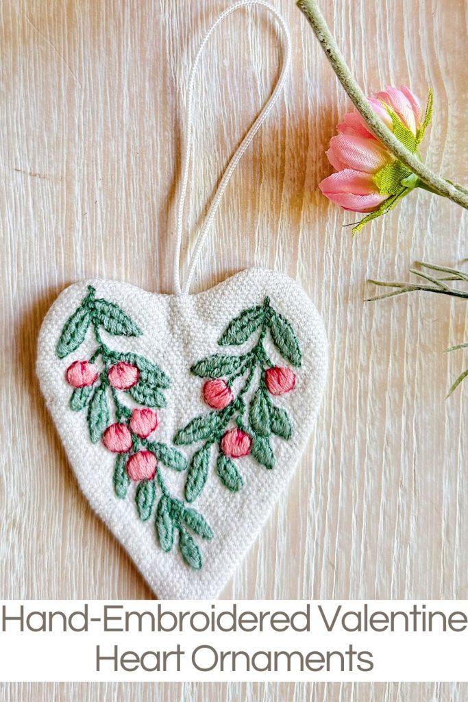 A heart-shaped ornament with hand-embroidered green leaves and pink berries, displayed next to a pink flower on a light wooden surface.
