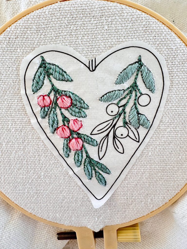 An embroidery hoop holds fabric with a heart-shaped outline, half stitched with green leaves and pink berries, the other half with unfinished green leaves and uncolored berries.