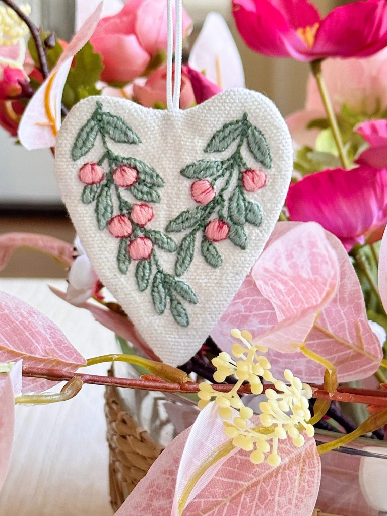 A white heart-shaped ornament with embroidered pink berries and green leaves hangs among pink flowers in a basket.
