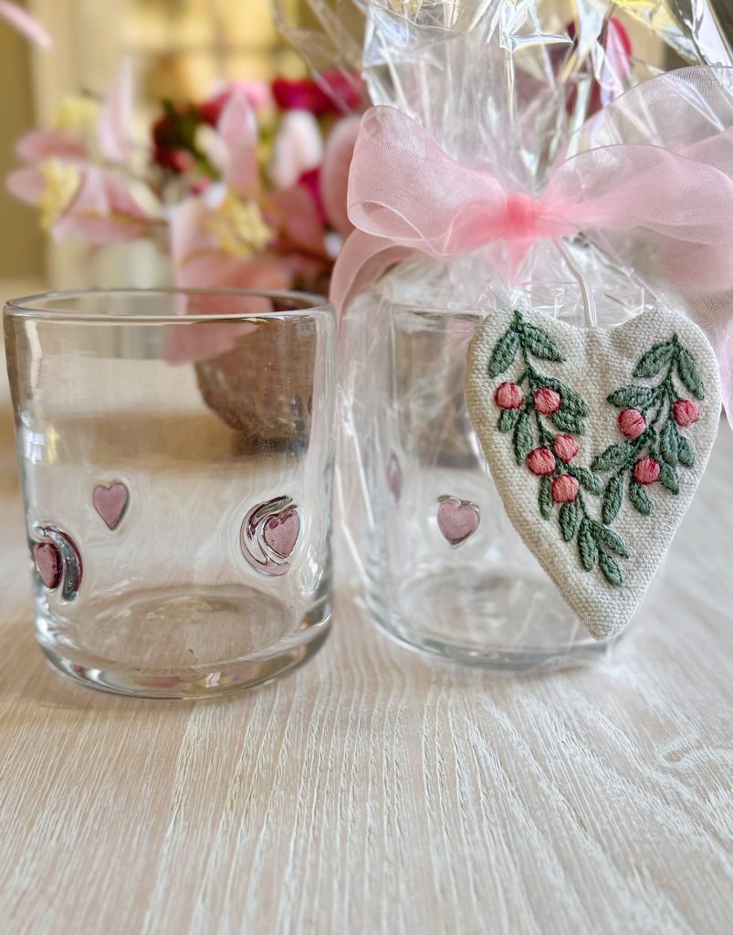 Two clear glass cups with pink heart decorations sit on a light wooden surface; one is wrapped in cellophane with a pink ribbon and a fabric heart ornament attached.