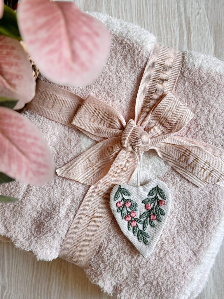 A folded pale pink towel tied with a beige ribbon and decorated with a heart-shaped ornament embroidered with flowers, next to pink leaves on a light surface.