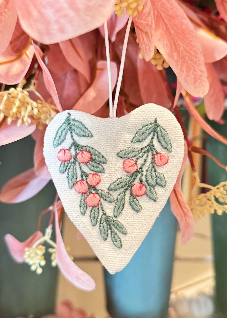 A white heart-shaped ornament with embroidered green leaves and pink berries hangs in front of pink flowers.