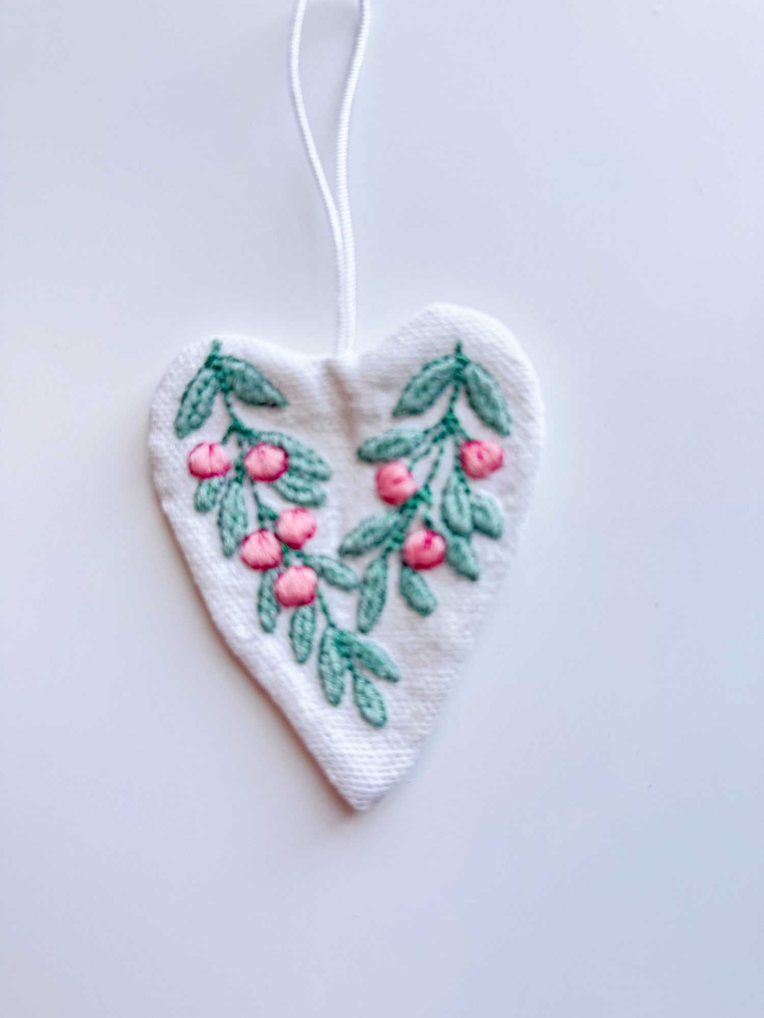 A white, heart-shaped fabric ornament with embroidered green leaves and pink berries, hanging by a white string against a plain background.