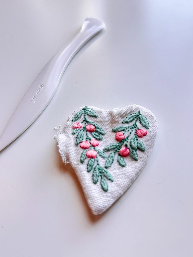 A white fabric heart with embroidered green leaves and pink flowers lies next to a white plastic tool on a white surface.