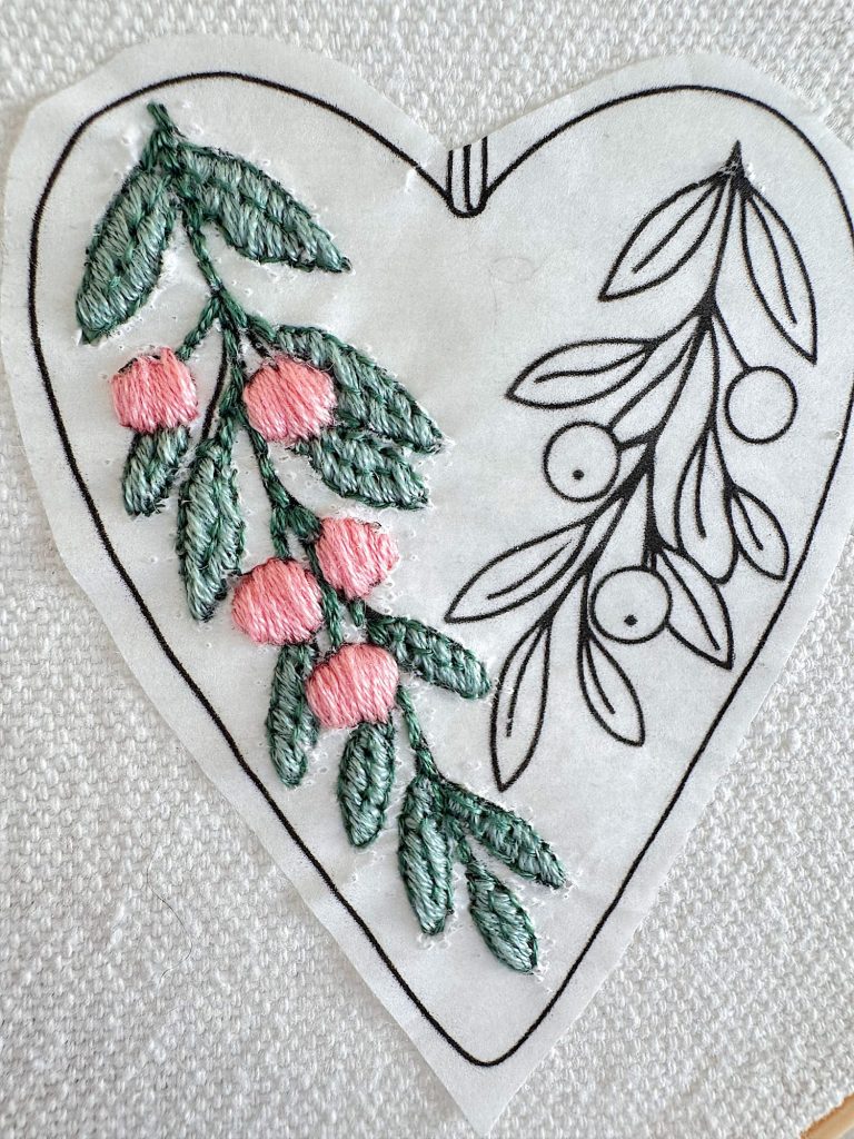 A heart-shaped embroidery pattern on fabric, partially stitched with green leaves and pink berries on one side, with the rest outlined in black.