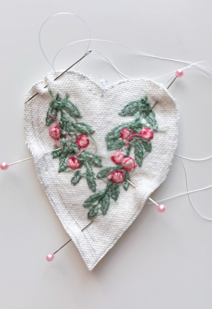 Heart-shaped piece of white fabric with embroidered pink flowers and green leaves, pinned to a surface with sewing pins and loose threads visible.
