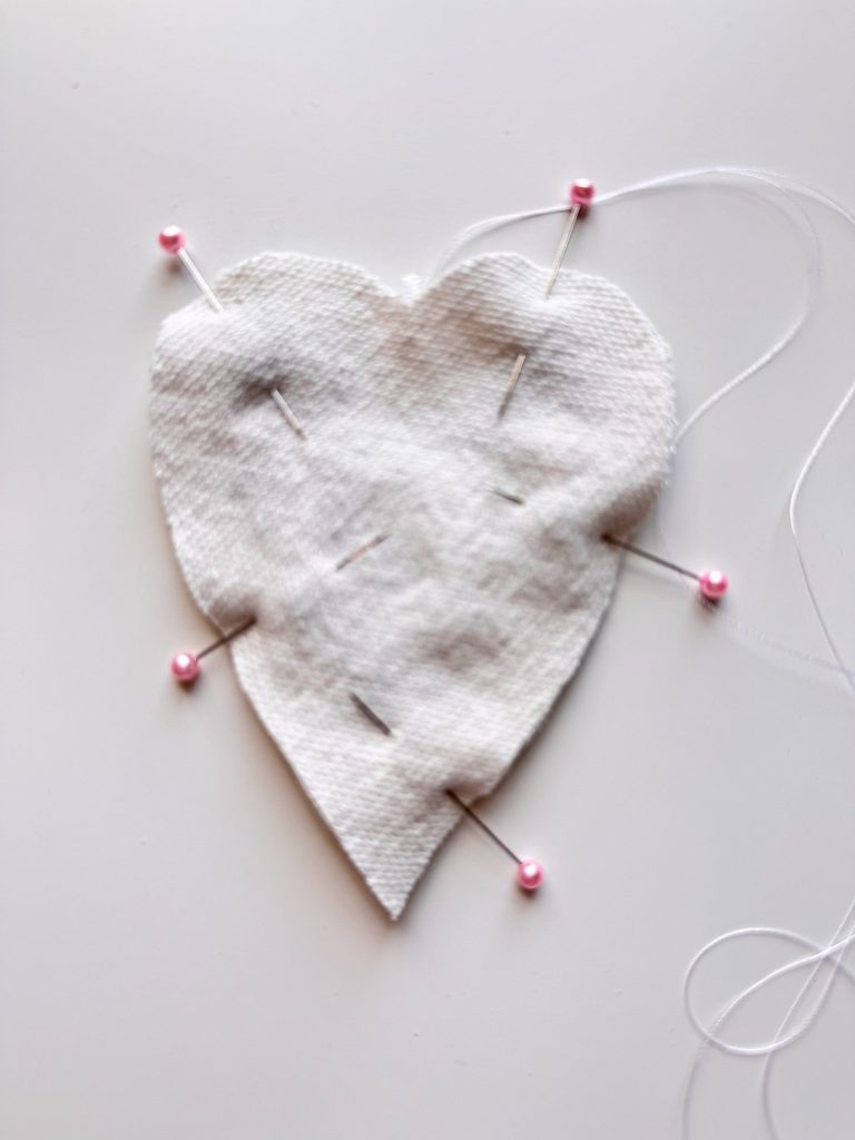 A piece of white fabric cut into a heart shape is pinned to a surface with five pink-headed sewing pins. White thread is visible on the right side.
