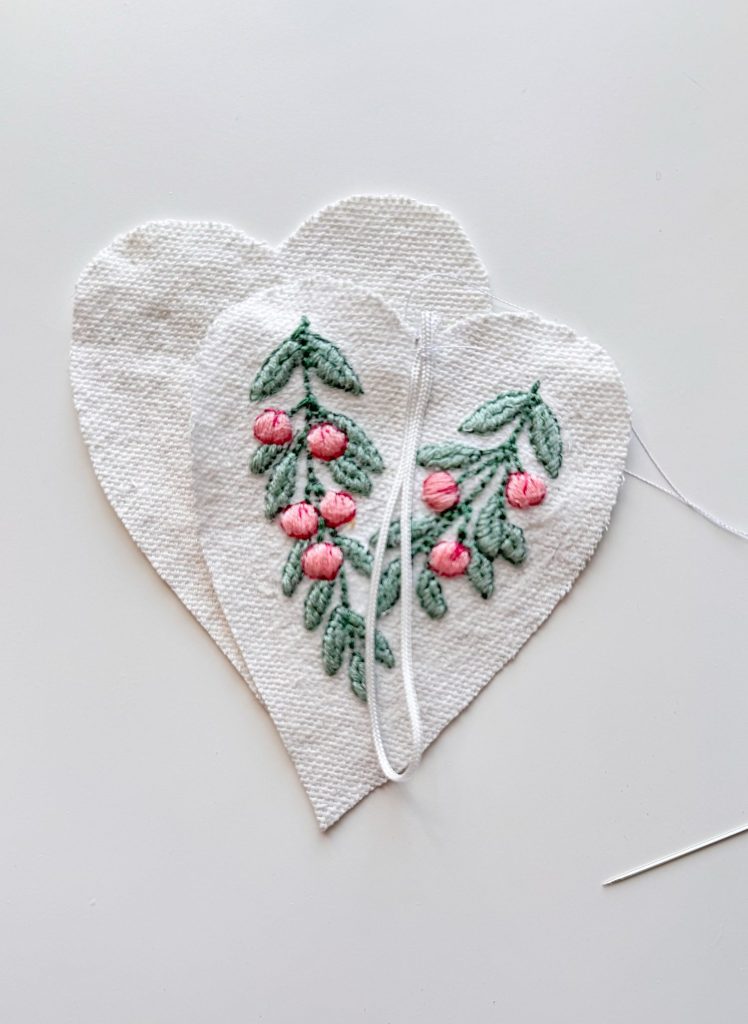 Two white fabric hearts; one is embroidered with green leaves and pink flowers, while the other is plain. A needle with white thread is placed nearby.