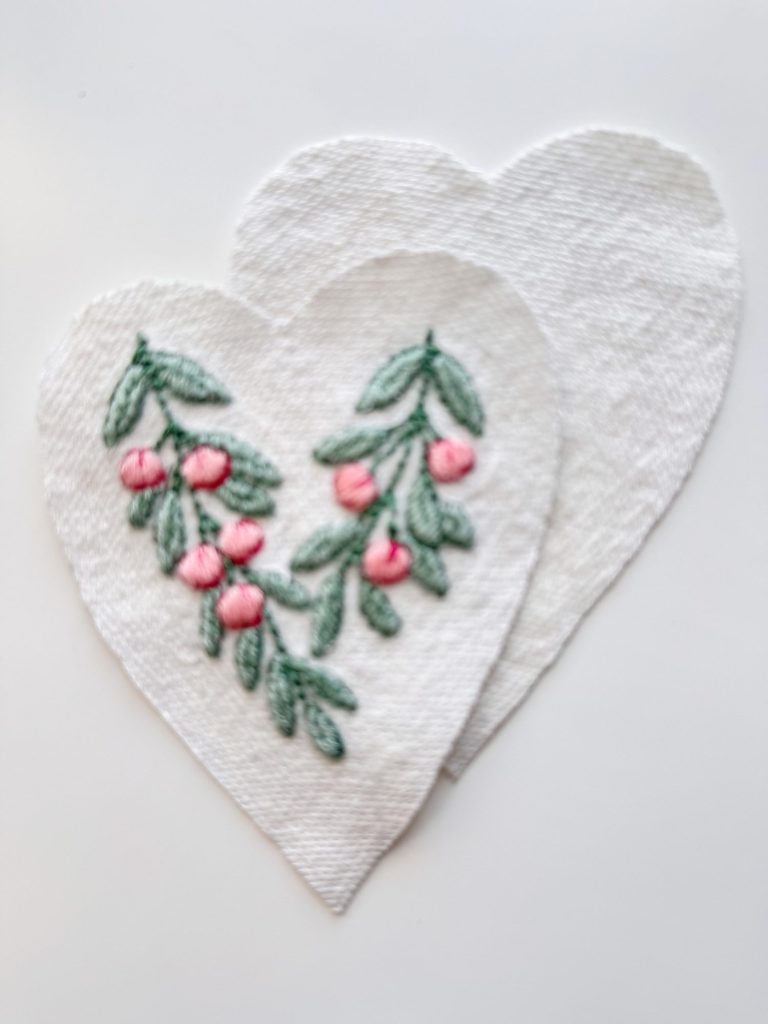 Two overlapping white fabric heart shapes, with one heart featuring embroidered green leaves and pink berries.