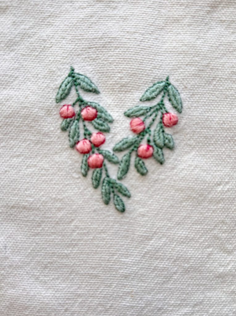 Embroidered design of green leaves and pink berries arranged in a V shape on a white fabric background.