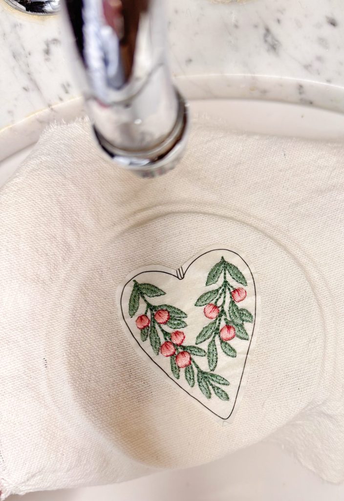 A white cloth with a heart-shaped design featuring green leaves and pink berries is placed under a glass plate near a metal faucet.