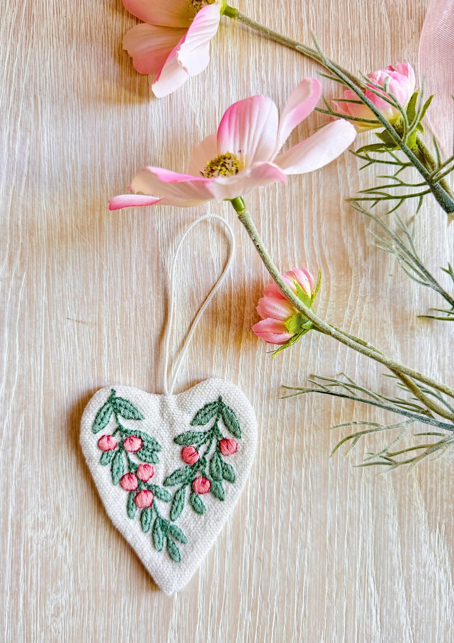 A heart-shaped fabric ornament with embroidered pink flowers and green leaves lies next to pink flowers on a light wooden surface.