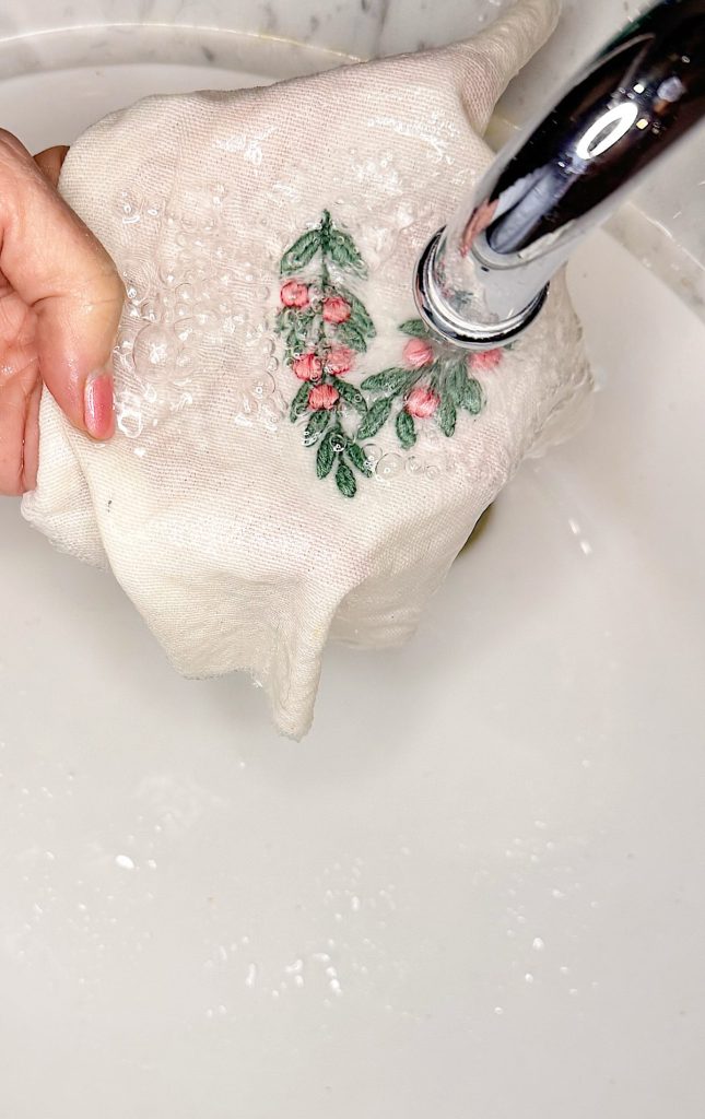 A hand washes a white cloth with pink and green floral embroidery under a running faucet in a sink.