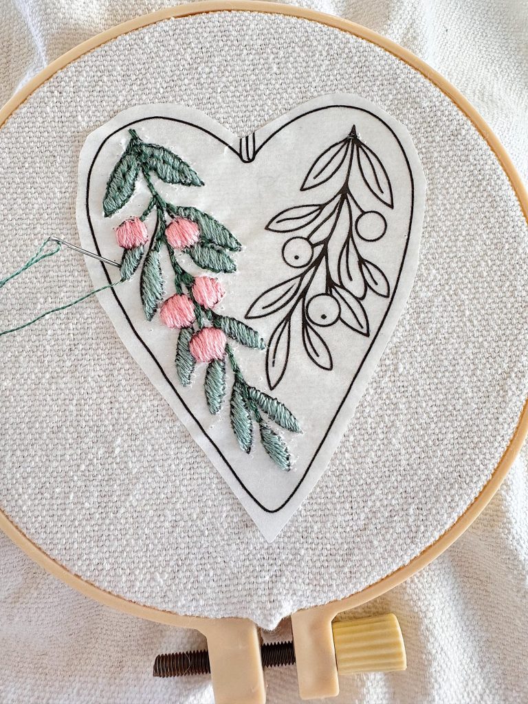 An embroidery hoop holds fabric with a heart-shaped floral design. Some leaves and berries are partially stitched with green and pink thread.