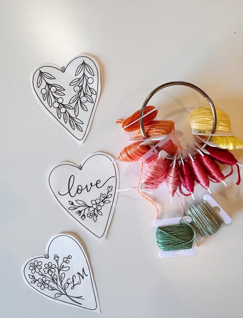 Three heart-shaped embroidery patterns on paper are next to a metal ring holding embroidery floss in various colors, including yellow, orange, pink, red, and green.