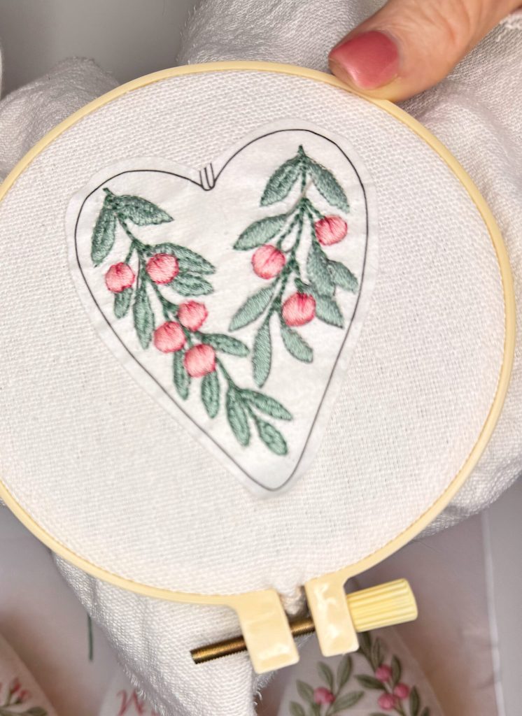 A hand holds an embroidery hoop with a heart-shaped pattern of green leaves and red berries stitched onto white fabric.