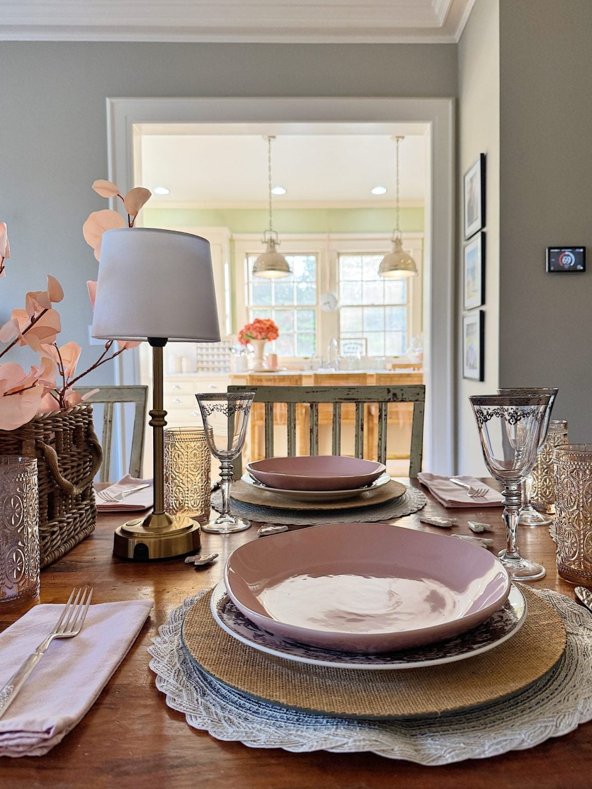 A dining table set for two with pink plates, wine glasses, a lamp, decorative flowers, and a view into a bright kitchen in the background.