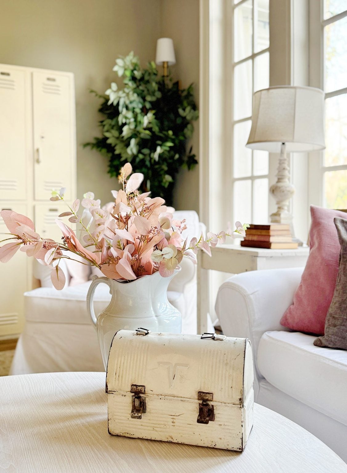 A white table with a vintage white metal box and a pitcher of pink flowers, set in a bright living room with white furniture, a lamp, and decorative lockers in the background.