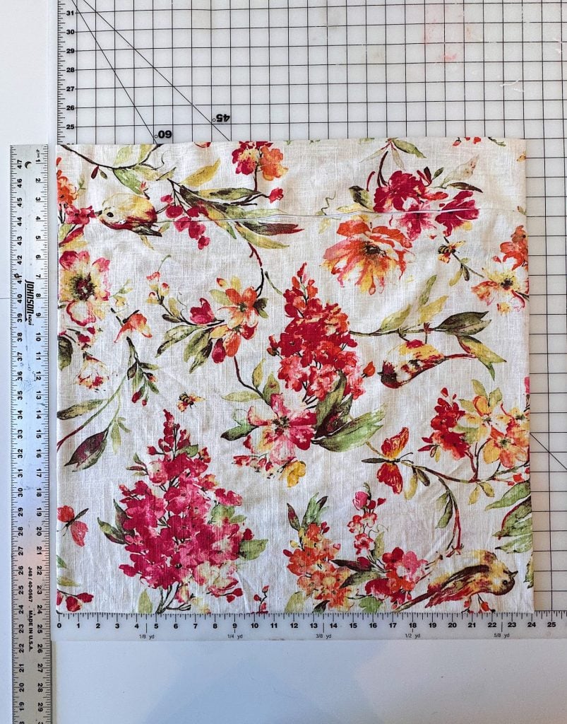 A square piece of white fabric with a red and green floral pattern is placed on a gridded cutting mat, with a metal ruler on the left and bottom edges for measurement.
