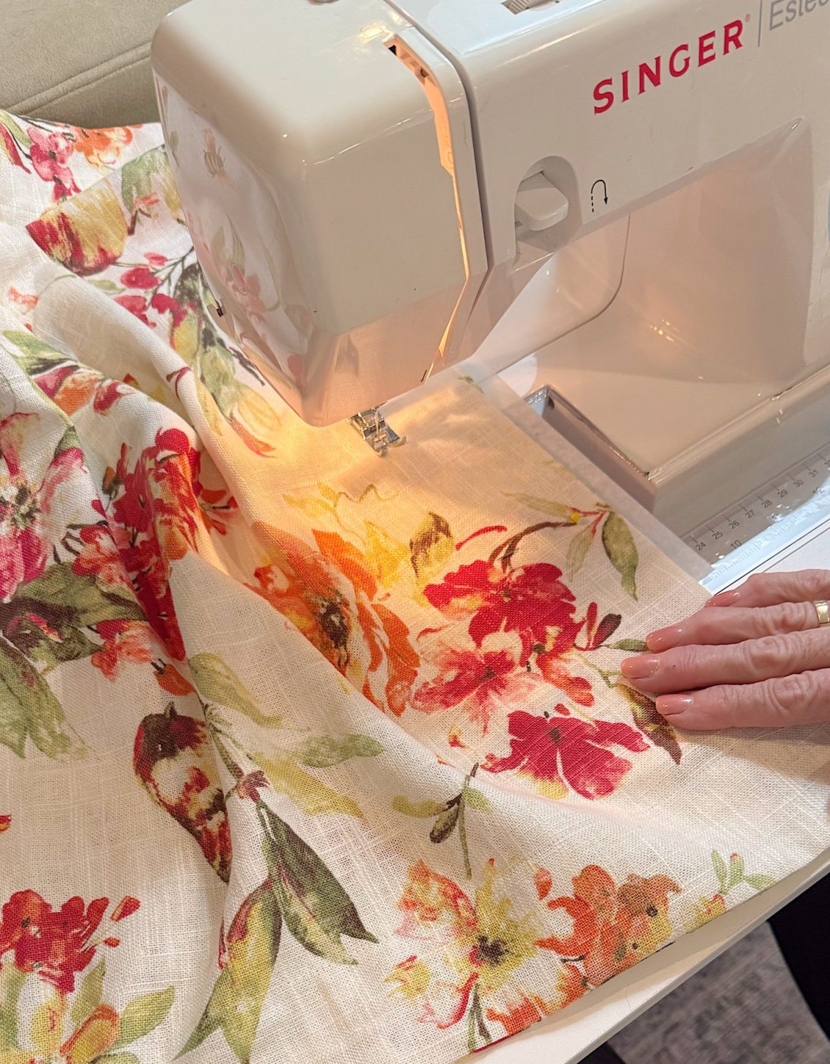 A sewing machine stitching floral-patterned fabric, guided by a person's hand.