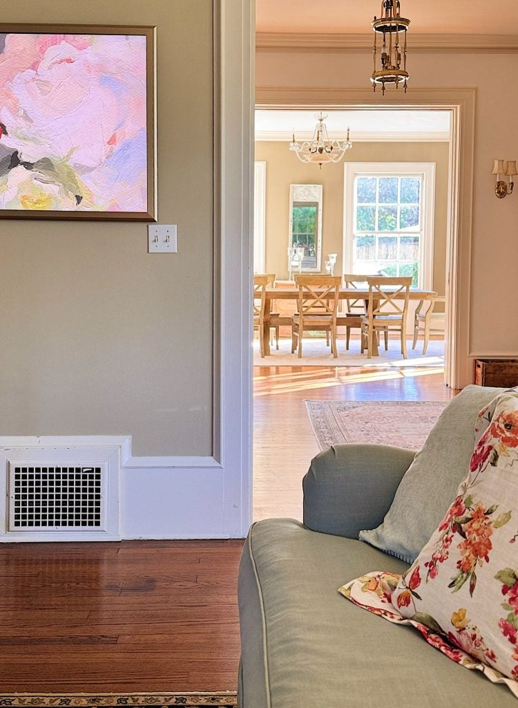 A living room with a floral pillow on a green couch, a vent and light switch on the wall, and a view into a bright dining room with wooden chairs and a chandelier.