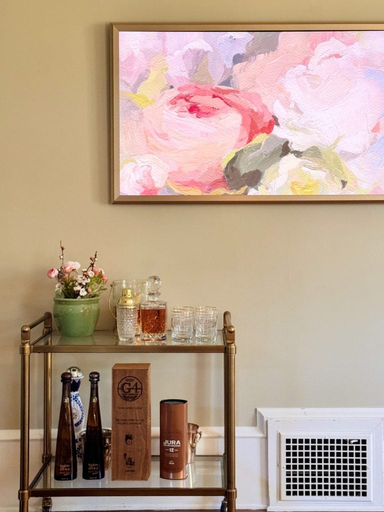 A bar cart with liquor bottles, glasses, a decanter, and a small flower arrangement below a framed floral painting on a beige wall.
