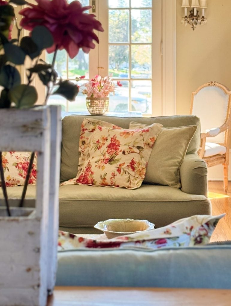 A sunlit living room with a green sofa, floral pillows, a white armchair, and a vase of pink flowers near glass doors.