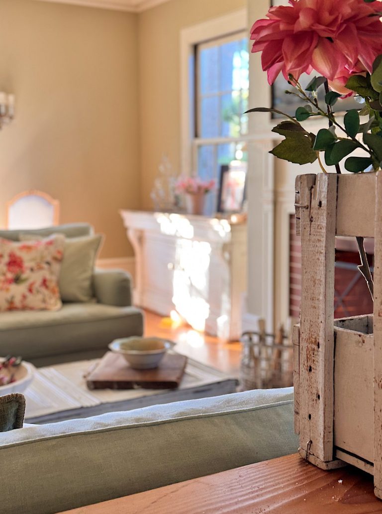 Sunlit living room with sage green sofa, floral cushions, a white coffee table, and a wooden box holding pink flowers in the foreground.