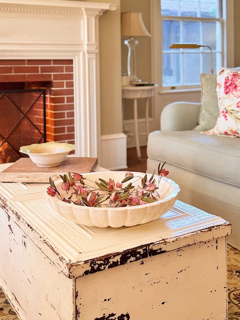 A white distressed coffee table holds a decorative bowl with pink flowers in a cozy living room with a lit fireplace, pale sofa, and floral pillow.