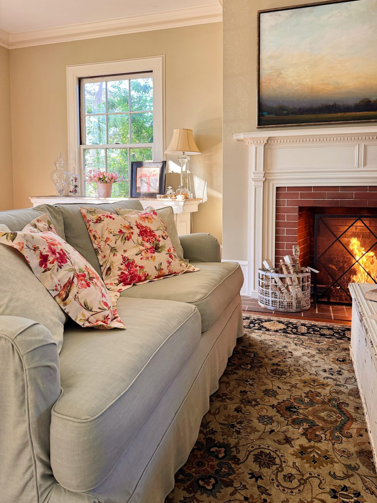 A cozy living room with a light-colored sofa, floral pillows, a lit fireplace, a patterned rug, and a window letting in natural light.