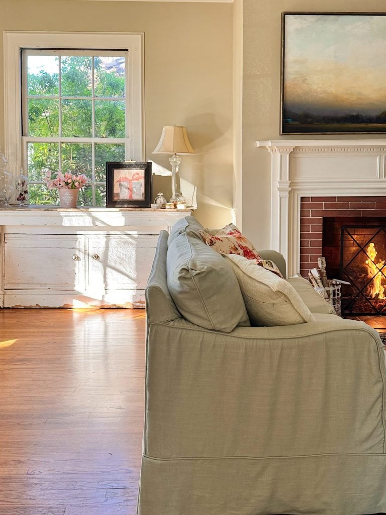 A cozy living room with a light gray sofa, a lit fireplace, a window letting in sunlight, and decorative items on a white cabinet.