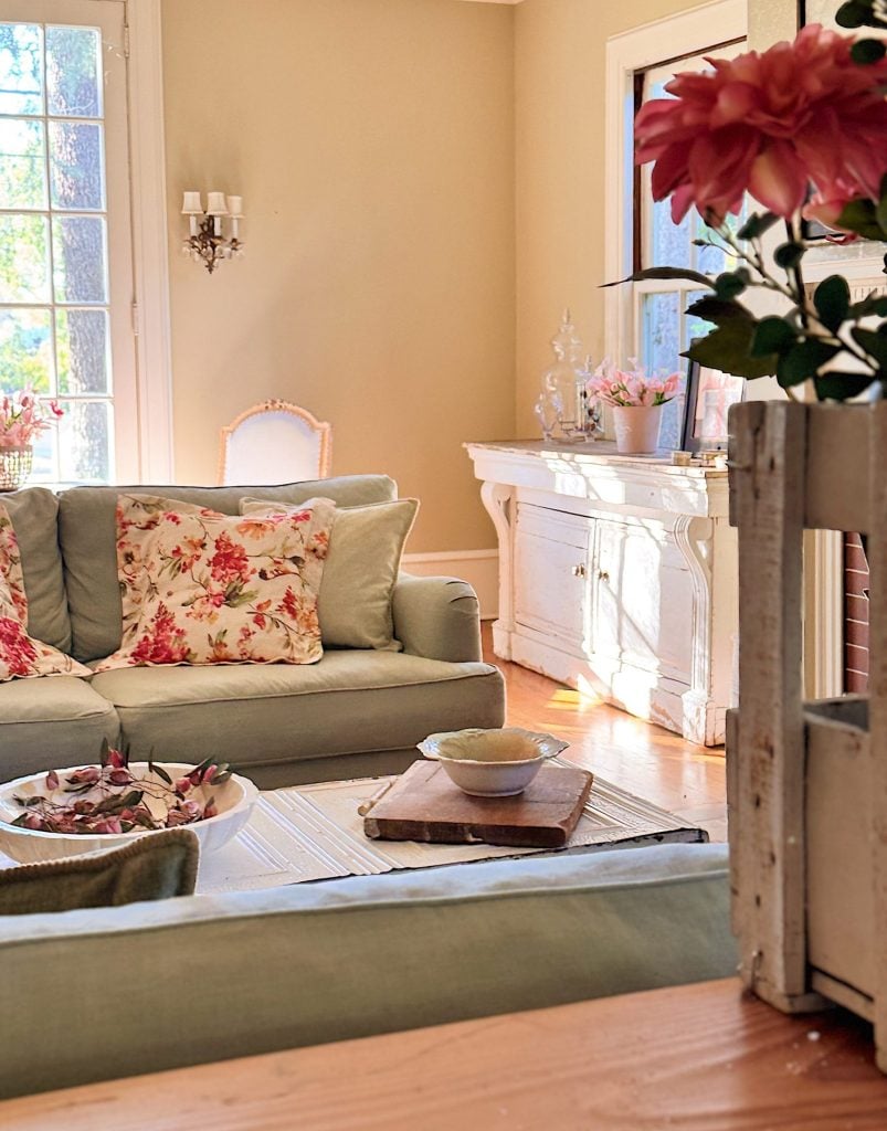 A sunlit living room with pastel green sofas, floral cushions, a white sideboard, and pink flowers in a vase; light streams in from tall windows.