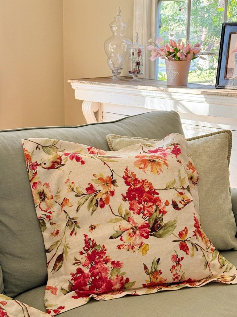 A floral-patterned pillow rests on a light green couch near a white mantel decorated with glass jars, a potted plant, and a framed picture by a sunlit window.