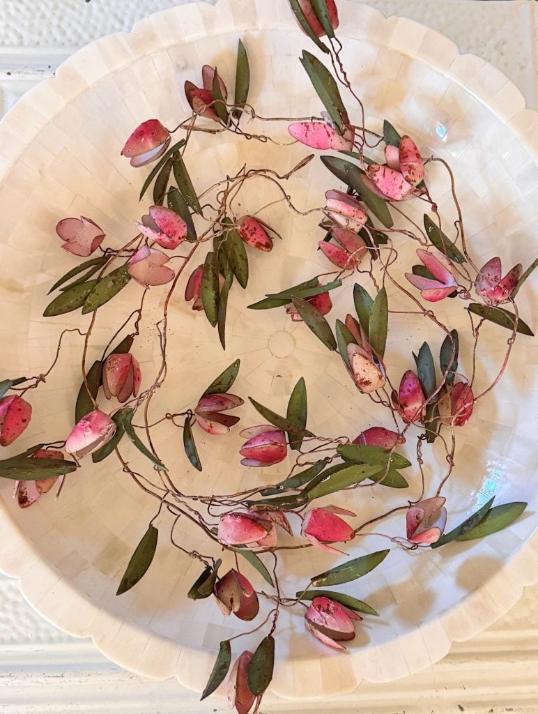 A strand of artificial vine with pink and green leaves is arranged in a loose spiral on a round, white, textured plate.