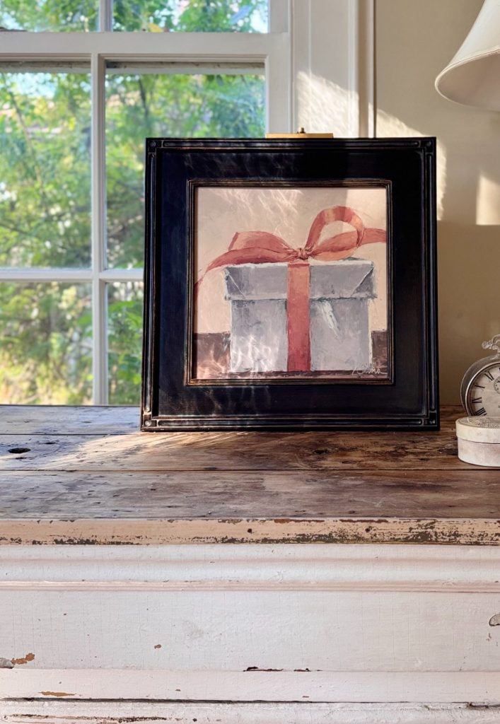 A framed painting of a silver gift box with a red ribbon sits on a rustic wooden table in front of a window.
