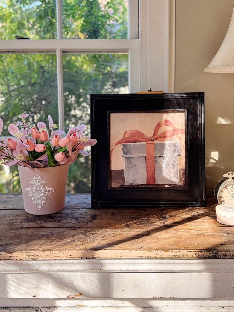 A framed painting of a gift with a red ribbon sits on a wooden table next to a pot of pink flowers, a small clock, and a lamp, with sunlight streaming through a window.