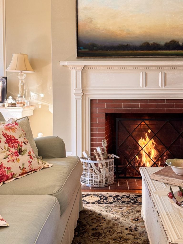 A cozy living room with a lit fireplace, a light-colored sofa with floral pillows, a patterned rug, and a coffee table. A lamp and decor items are on a side table and mantel.