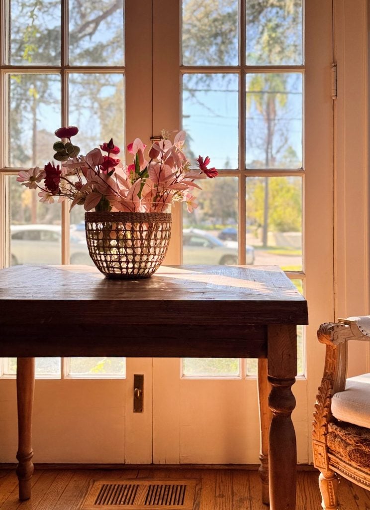 A vase with pink flowers sits on a wooden table in front of glass doors, with sunlight streaming in and a view of trees and parked cars outside.