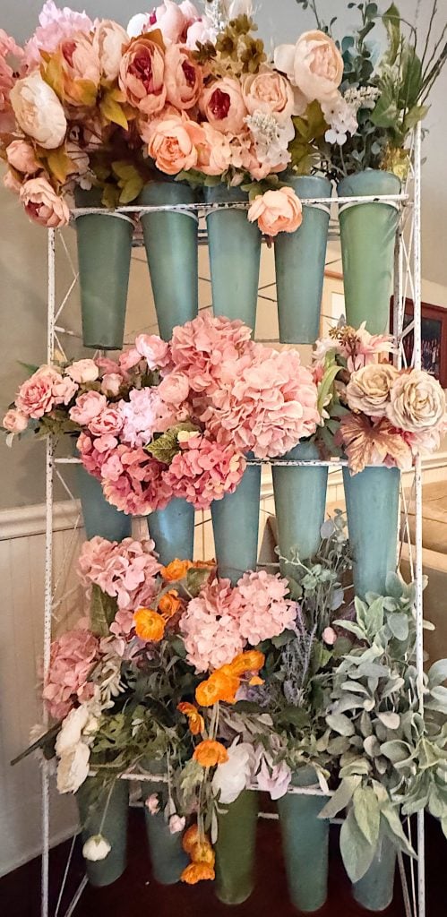 A metal rack holds rows of teal vases filled with various pink, peach, and orange artificial flowers and green foliage.