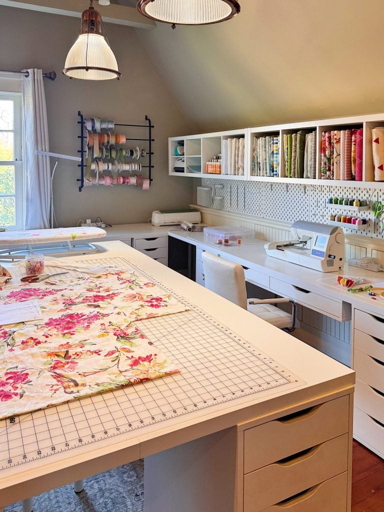 A well-organized sewing room with a large worktable, sewing machine, fabric rolls, ribbon rack, storage shelves, and natural light from a window.