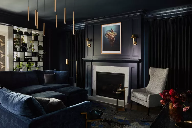 A modern living room with dark walls, a blue sectional sofa, a fireplace, an armchair, hanging lights, and a framed artwork above the mantel.