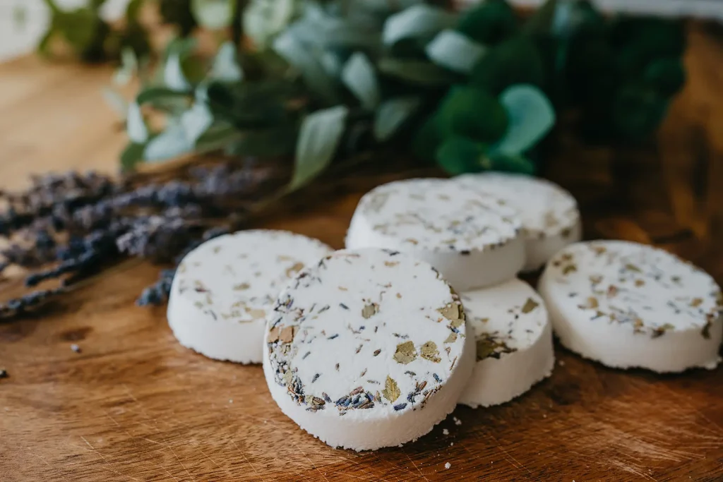Five round, white bath bombs with dried herbs on top are arranged on a wooden surface, with green leaves and dried lavender in the background.
