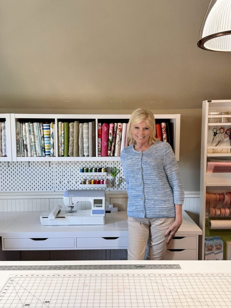 A woman stands in a sewing room with fabric neatly organized on shelves, thread and scissors displayed, and a sewing machine on the table.