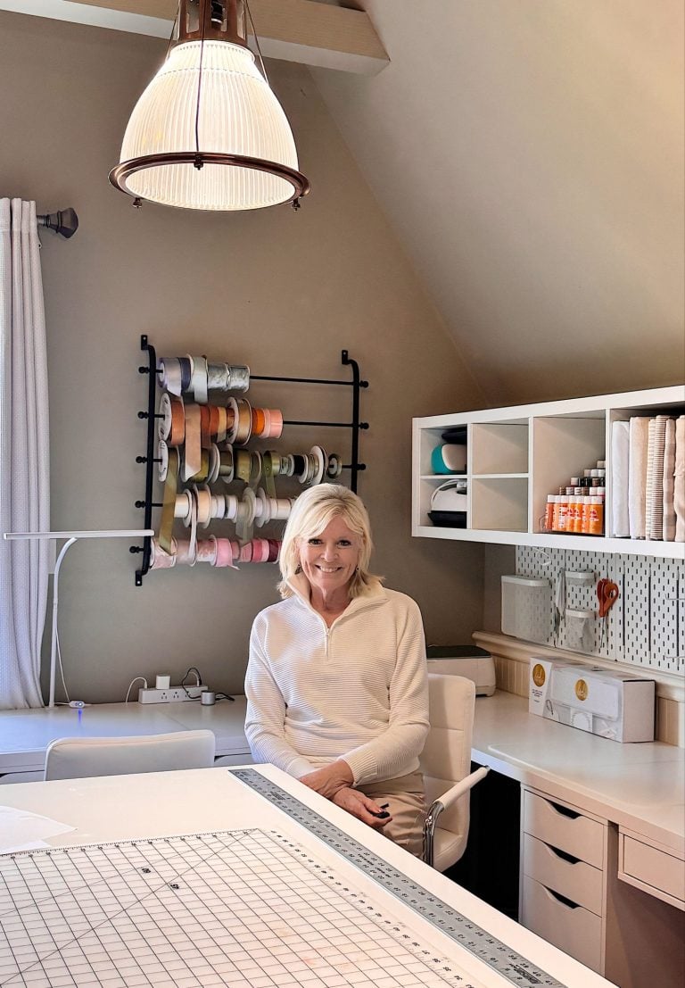 A woman sits at a desk in a craft room with organized shelves, ribbon spools on the wall, and a large cutting mat on the table.
