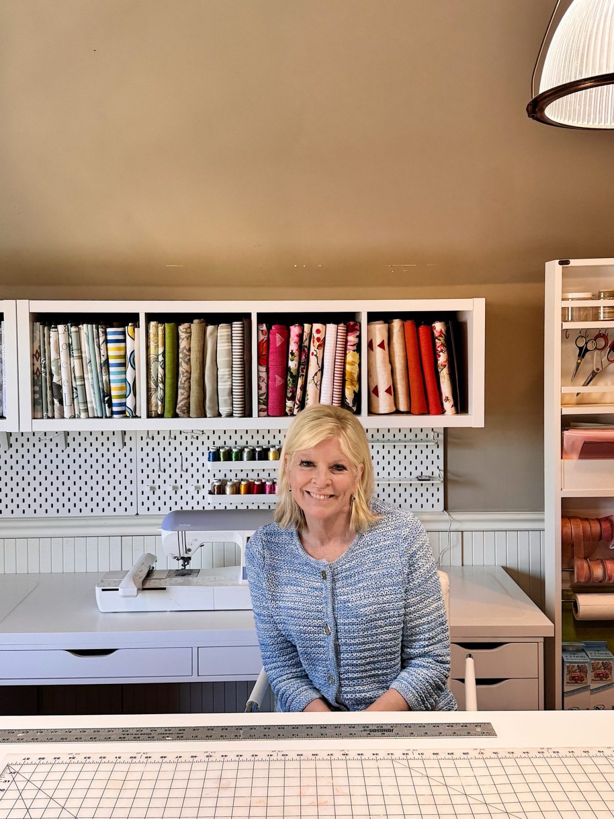 A woman sits at a white desk in a sewing room, with fabric, ribbons, and sewing supplies organized on shelves and a sewing machine behind her.