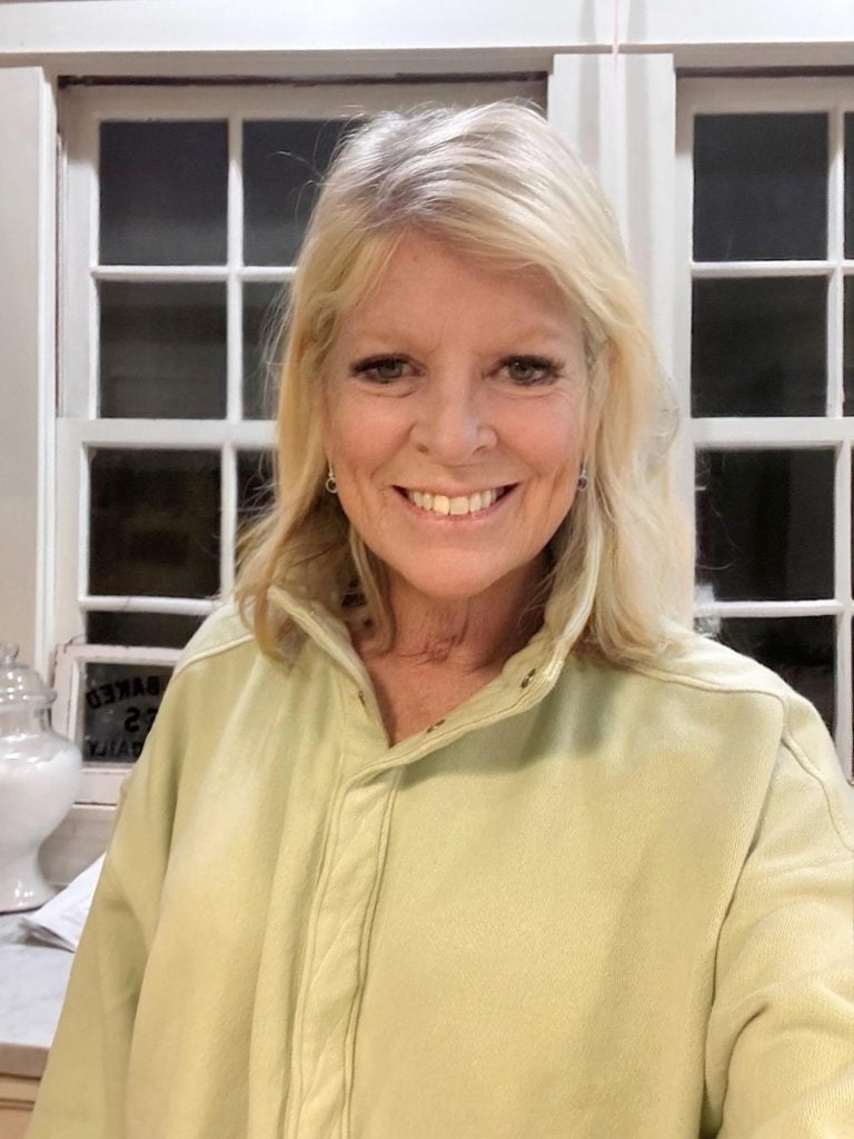 A woman with blonde hair wearing a light green collared shirt smiles while standing indoors in front of a window.