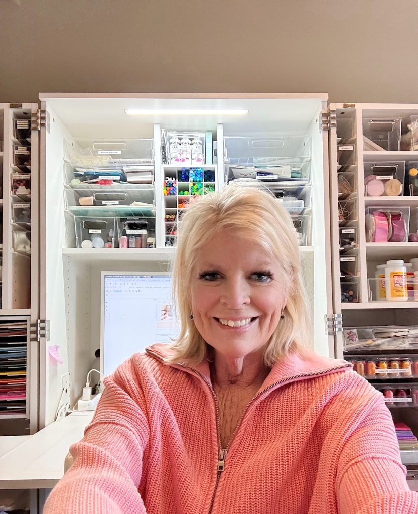 Woman in a pink zip-up sweater smiles while taking a selfie in front of an organized craft workspace with shelves and storage bins.