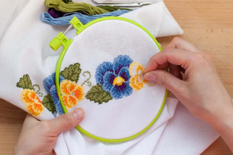 Hands embroidering a floral cross-stitch pattern with yellow, blue, and green thread on white fabric held in a green embroidery hoop.