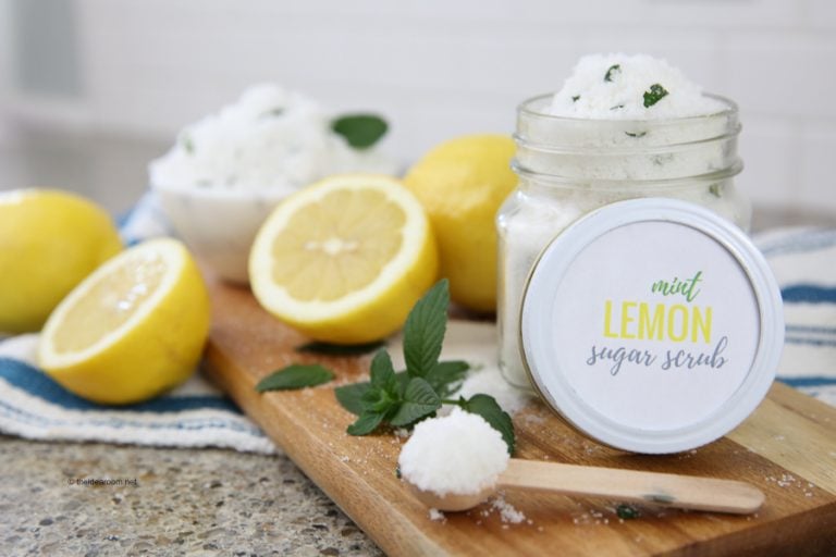 A jar labeled "mint lemon sugar scrub" sits on a cutting board with sliced lemons, mint leaves, and a small wooden spoon filled with scrub.