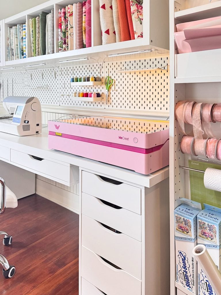 A craft room with shelves of fabric, a sewing machine, thread spools, a pink cutting machine, and organized storage drawers on a wood floor.
